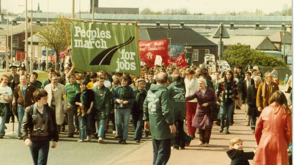 A group of people on a march through an industrial areas. They are holding a large banners, one reads 'People's March for Jobs' 