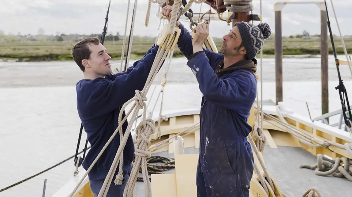 two people pulling a rope on a historic ship, with a river estuary in the background