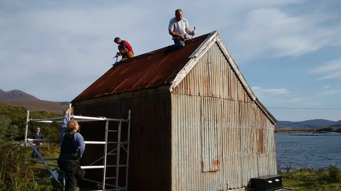 Men working on a small corrugated building with water and mountains in the distance.