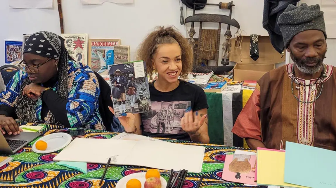 Three black people take part in a workshop on black history. All three people have dark skin of various shades, the focus of the image is a woman holding a magazine with black people on the cover