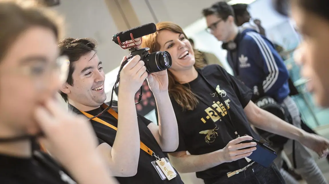 Two people wearing Reimagine, Remake, Replay tshirts using filming equipment at an event