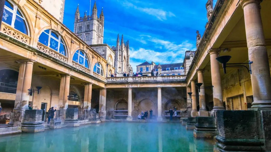 Looking across the water at the Roman Baths in Bath, people standing on the edges and looking down from a walkway above with