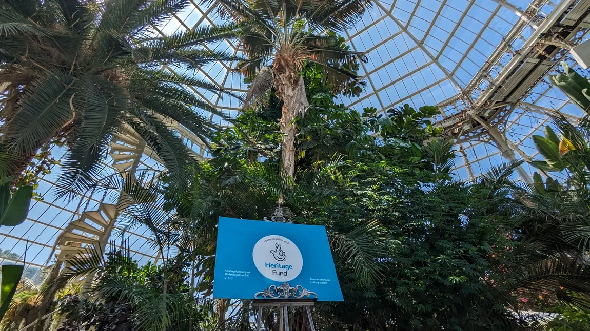 Palm trees and plants inside a glassed roofed building. A board with the Heritage Fund logo is on display on an easel. 