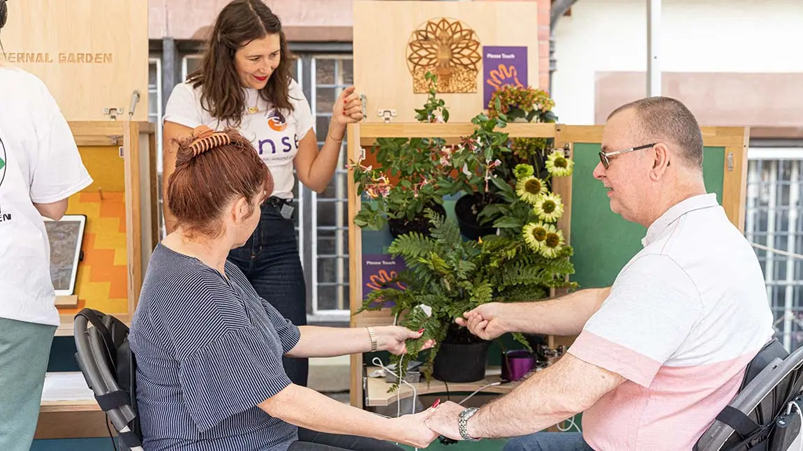 Two people sit in chairs holding hands with their other hands touching the leaves of a potted plant. Wires run from the plant to pads on the people's wrists. A smiling person in a Sense t-shirt watches.