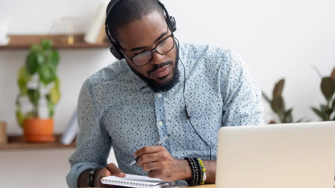 A black man waering headphones, looking at a laptop computer and taking notes in a notebook