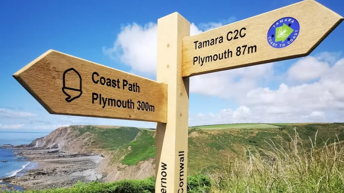 A wooden signpost pointing one way to Plymouth 300 miles away, and another way to the Tamara Coast to Coast route with Plymouth 87 miles away.