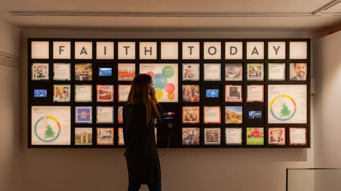 A visitor listens through an exhibition headset in front of a display of information about faith
