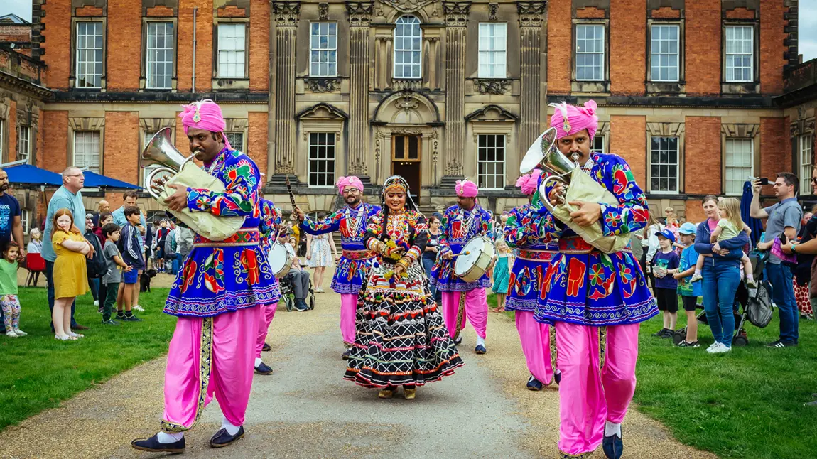 An Asian brass band, with musicians and dancers dressed in colourful clothes, walking down a path in front of a stately home