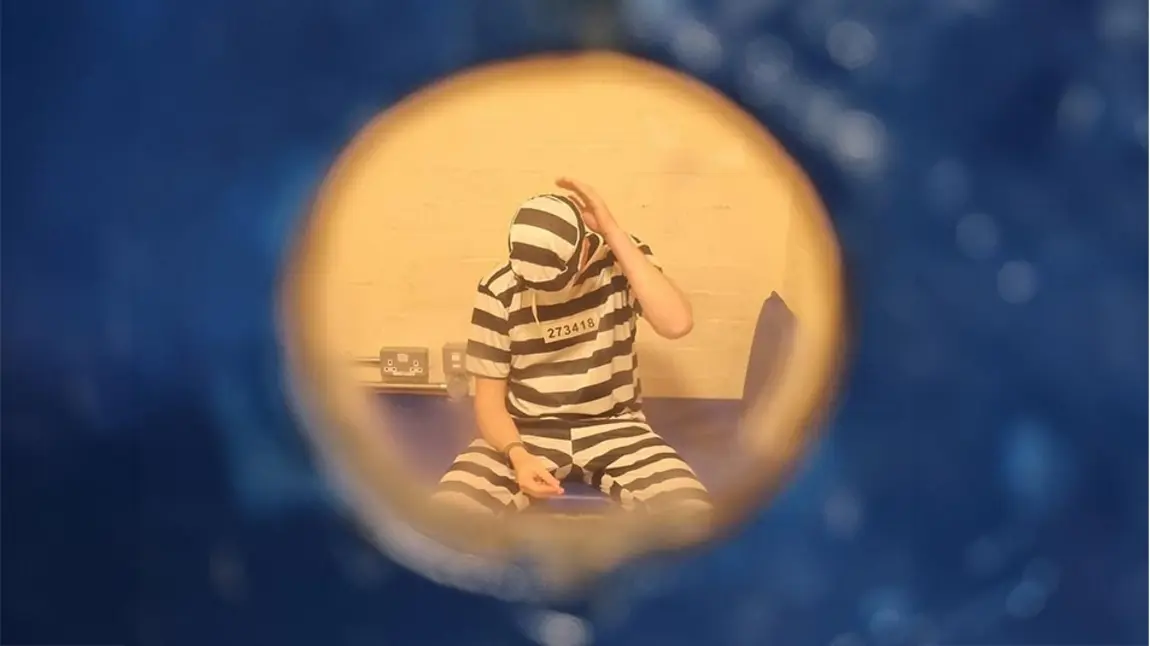A photo taken through a keyhole of a prisoner in a cell, wearing striped black and white clothing.