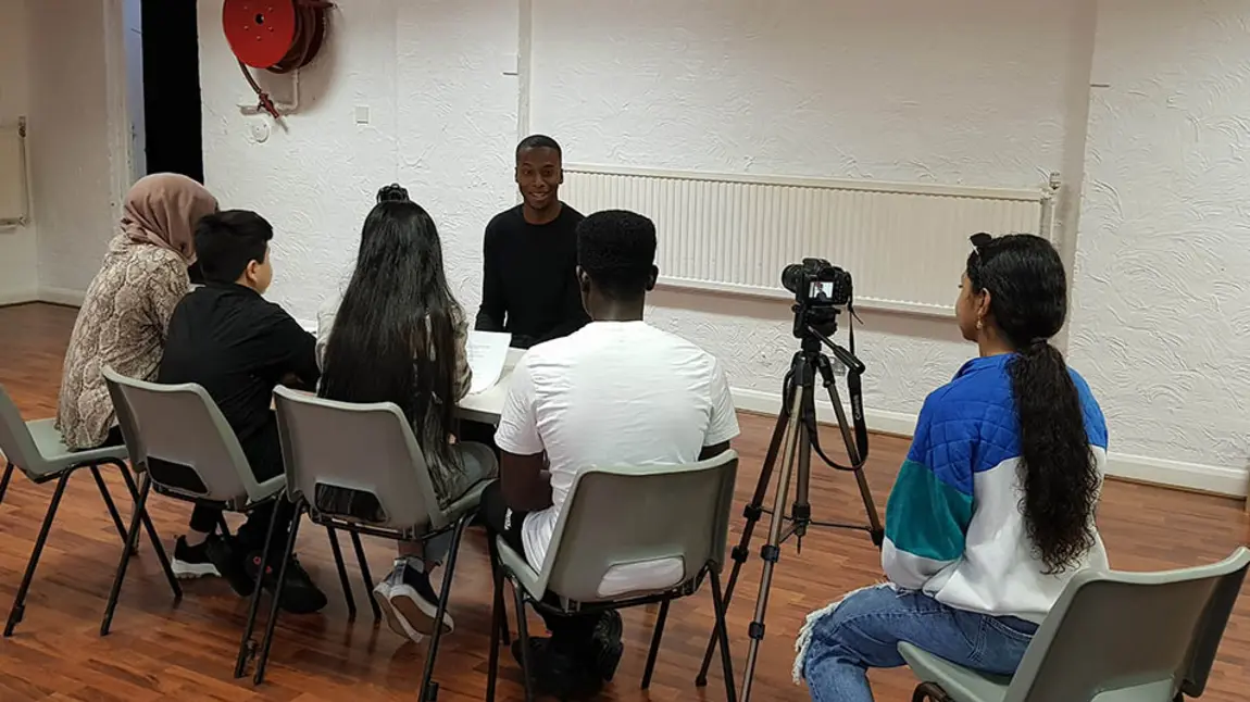 A group of young people sit a table interviewing another young person, with another person to the side operating a camerawith 