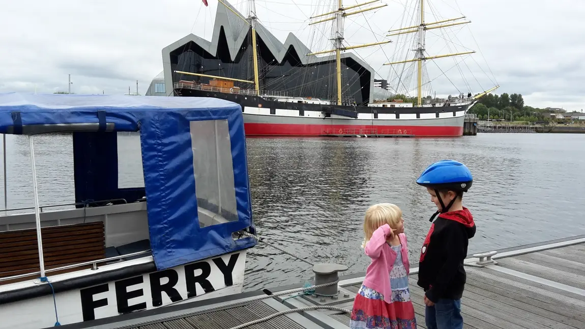 A boy and a girl, stand facing each other on a waterfront near Glasgows Riverside Museum