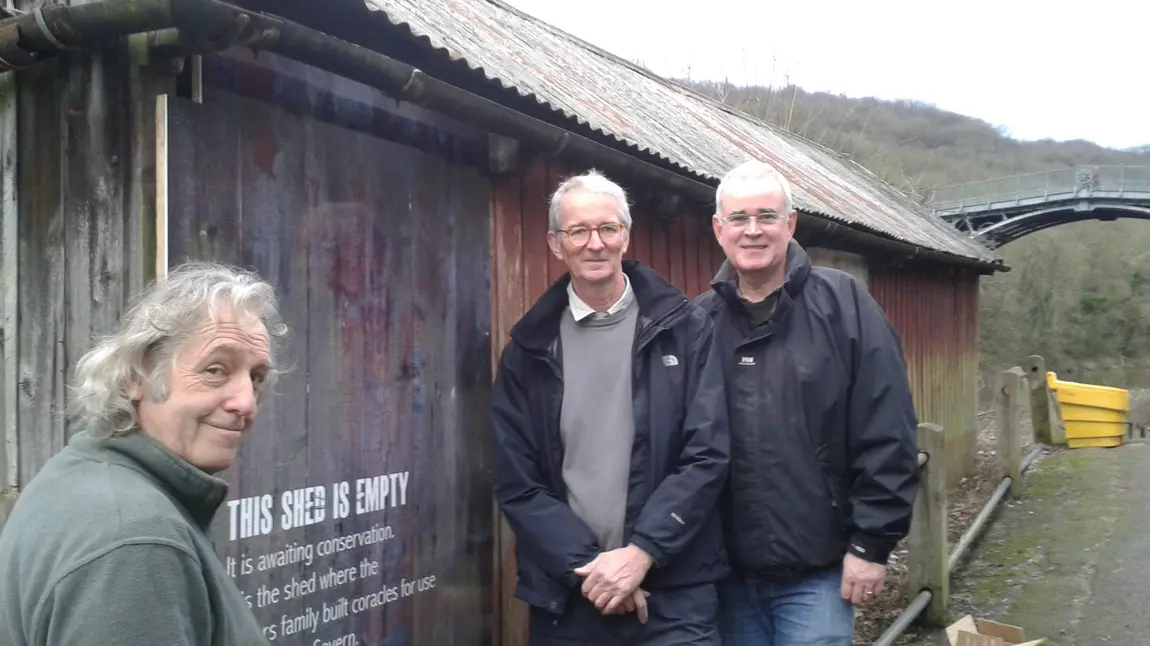 Three people stand outside a large closed shed with a sign saying awaiting restoration