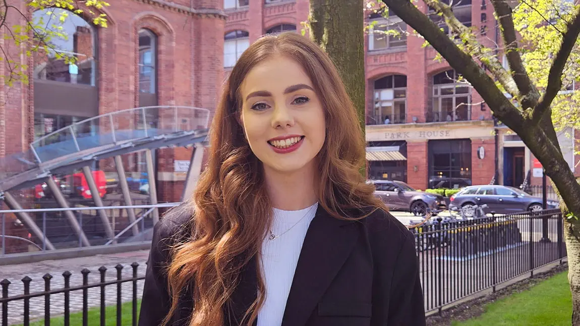 A young white woman with long red-blonde hair wearing a white top and black jacket standing in an urban area.