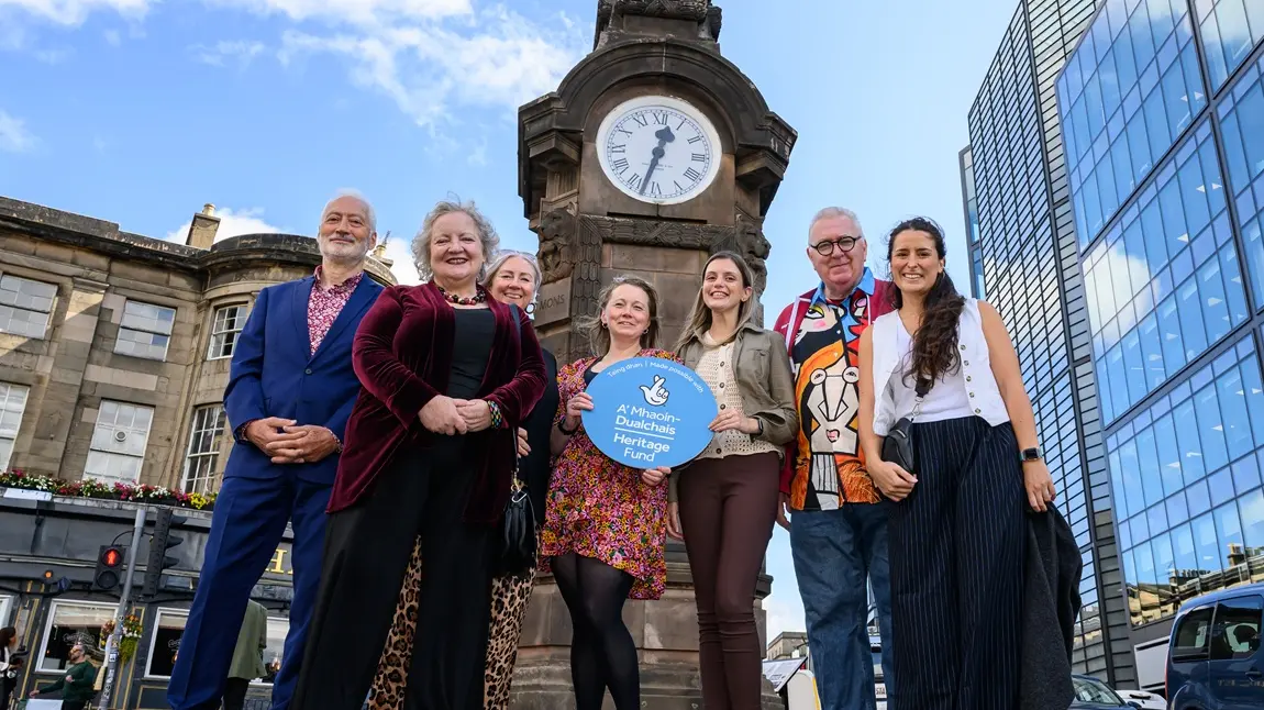 A project group of seven people pose for a photograph in front of the Hear of Midlothian Memorial Clock. Karen Mailley stands at the centre of the group, holding a Heritage Fund branded roundel.