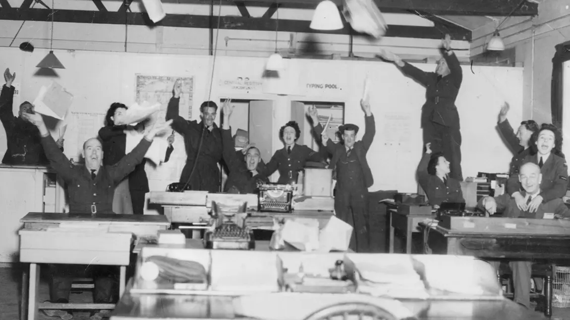 A black and white photograph showing RAF staff celebrating in an office, throwing their arms and papers in the air.