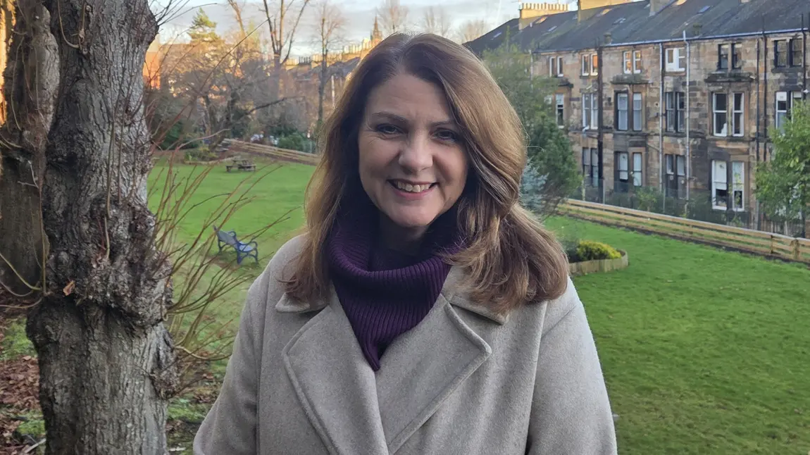 Heather Reid, wearing a grey woollen coat and purple scarf, stands smiling in a field behind a row of Scottish tenement houses.