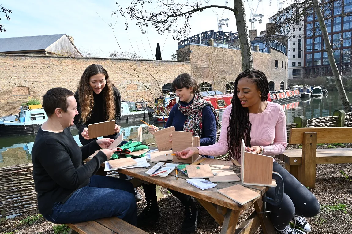 A group of people sitting next to a canal and building wooden bird boxes on a picnic bench