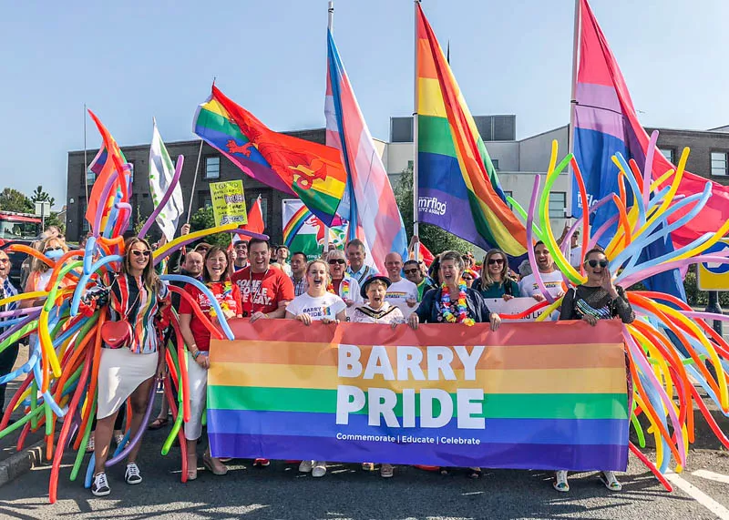 A crowd with colourful balloons and flags holding a rainbow pride banner that reads Barry Pride, 