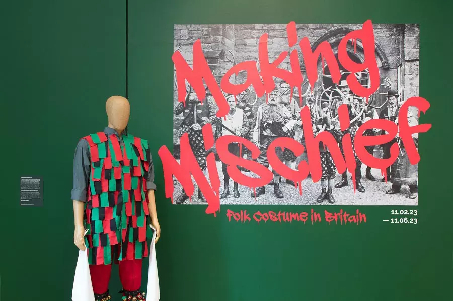 A mannequin wearing a bright green and black folk costume standing in front of a sign that reads 'Made in Mischief, Folk Costume in Britain'