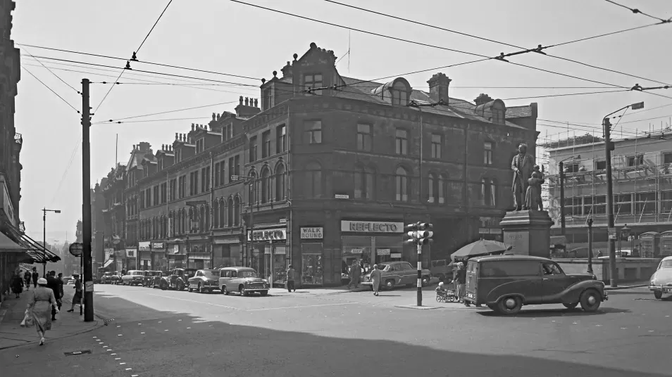 An old black and white photograph of a busy town centre with shops, cars and people walking