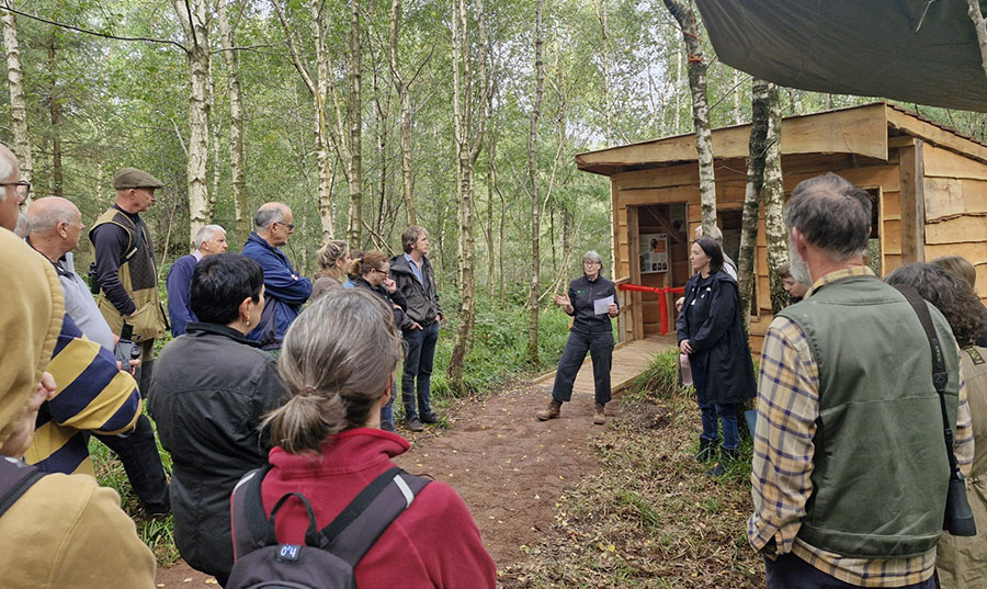 A group of people listen to a speech in front of a wooden hut in a forest